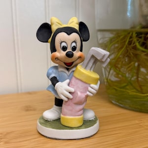 May include: A Minnie Mouse figurine in a light blue dress and white shoes, holding a pink and yellow golf club bag. The figurine has a yellow bow and is standing on a green and white base.