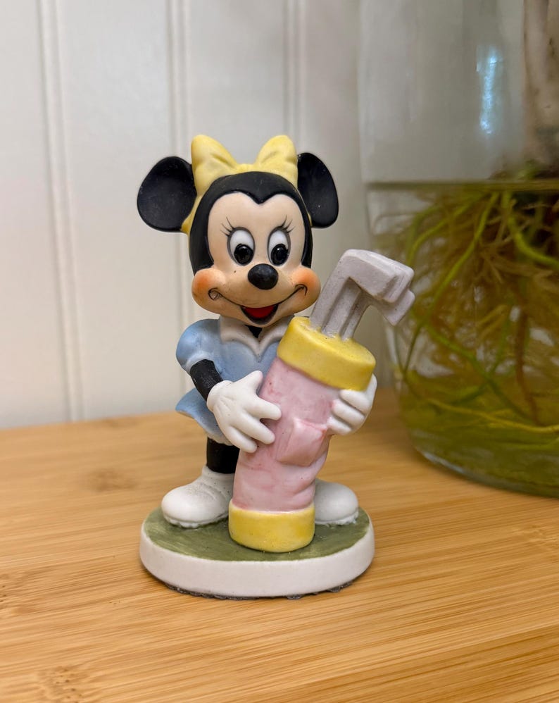 May include: A Minnie Mouse figurine in a light blue dress and white shoes, holding a pink and yellow golf club bag. The figurine has a yellow bow and is standing on a green and white base.