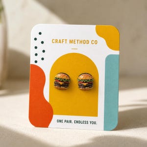 May include: A pair of miniature hamburger stud earrings featuring detailed layers of bun, lettuce, tomato, cheese, and patty. The earrings are attached to a white display card with the text CRAFT METHOD CO and ONE PAIR. ENDLESS YOU.