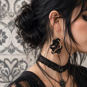 May include: A dangling earring featuring a black coiled snake design encased within a circular gold-toned frame. The earring hangs from a thin silver-colored hook, showcasing a detailed, textured serpent motif that creates a bold, dark aesthetic.
