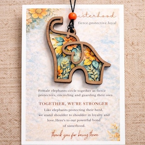 May include: Wooden elephant pendant with floral design, hanging from a black cord. The pendant is on a card with the words "sisterhood" and "fierce.protective.loyal". The card also includes text about sisterhood and loyalty.
