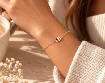 May include: A delicate gold chain bracelet featuring two small charms: one red teardrop-shaped stone and one clear rectangular baguette-cut stone. The bracelet is secured with a thin gold-toned metal chain that rests against the wrist.