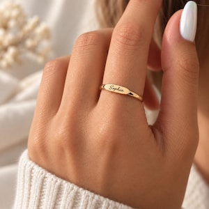 May include: A polished gold-tone ring featuring a slim, rounded band and a central horizontal bar plate. The plate is engraved with the name Sophia in a black, cursive script font. The ring has a smooth, reflective metallic finish throughout.