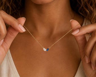 May include: A delicate gold chain necklace featuring two small, marquise-cut gemstones positioned at the centre. One stone is clear and colourless, while the other is a deep, vibrant blue. The necklace is held by two hands, showcasing the pendant.