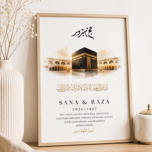 May include: A framed vertical print featuring a watercolour illustration of the Kaaba in Mecca. The design includes Arabic calligraphy, the names SANA & RAZA, the years 2026 and 1447, and English text that reads: MAY ALLAH ACCEPT YOUR HAJJ.
