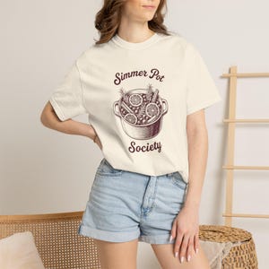 May include: A cream-coloured short-sleeve t-shirt featuring a dark brown graphic of a pot filled with citrus slices, berries, and herbs. The text "Simmer Pot Society" is printed in dark brown above and below the illustration of the pot.