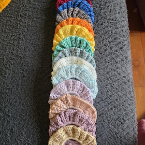 May include: A collection of colorful, crocheted scrunchies in various shades, including black, red, blue, gray, orange, yellow, green, white, and purple. The scrunchies are arranged in a vertical stack, showcasing their textured, ruffled design. These hair accessories are perfect for adding a touch of handmade charm.