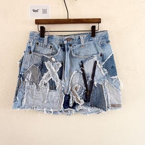 May include: Light blue denim skirt with patchwork design. The skirt features a frayed hem and various denim patches sewn together. The skirt is hanging on a wooden hanger against a neutral background. The skirt has a zipper and button closure.