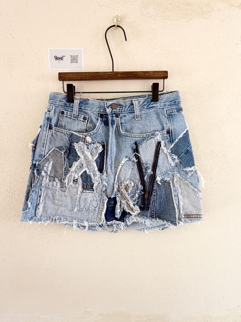 May include: Light blue denim skirt with patchwork design. The skirt features a frayed hem and various denim patches sewn together. The skirt is hanging on a wooden hanger against a neutral background. The skirt has a zipper and button closure.