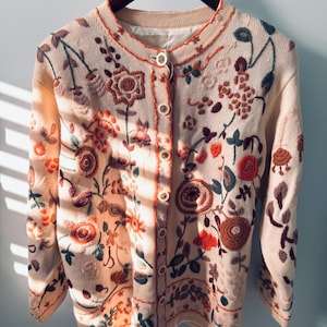 Könnte beinhalten: Cremefarbene Strickjacke mit aufwendiger Blumenstickerei in Rot-, Orange-, Braun- und Grüntönen. Die Strickjacke hat einen runden Ausschnitt, eine Knopfleiste vorne und lange Ärmel. Das florale Design bedeckt die Vorderseite und die Ärmel.