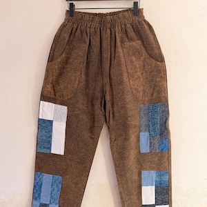 Peut inclure: Pantalon marron avec de grandes poches avant et des détails de patchwork. Les patchs sont des carrés de tissus bleus, blancs et denim. Le pantalon est suspendu à un cintre en bois.