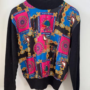 Peut inclure: Un pull noir vintage avec un col montant et des manches longues. Le devant présente un motif coloré avec des accents roses, bleus et dorés. Le motif comprend des images de roues, de chapeaux et d'autres éléments décoratifs.
