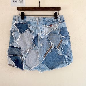 May include: A denim mini skirt with a patchwork design. The skirt is made from various shades of blue denim, with visible stitching and frayed edges. The skirt is hanging on a wooden hanger.