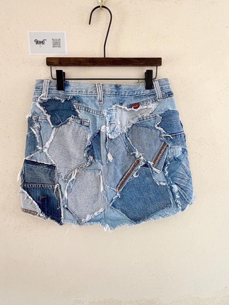 May include: A denim mini skirt with a patchwork design. The skirt is made from various shades of blue denim, with visible stitching and frayed edges. The skirt is hanging on a wooden hanger.
