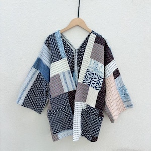 May include: A patchwork jacket with a V-neck design, featuring a variety of patterned fabric squares in shades of blue, gray, and brown. The jacket is hanging on a wooden hanger against a white wall. The sleeves are three-quarter length.