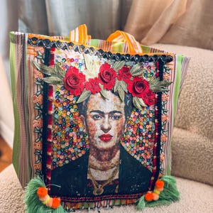 May include: A vibrant tote bag with a Frida Kahlo portrait. The bag features a green and white striped design, red rose embellishments, and green tassels. The Frida image is framed by colourful sequins and embroidery.