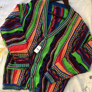 May include: A vibrant, vintage cardigan in a striped design, featuring green, blue, orange, and red. The button-up sweater has pockets and a tag reading "boho". It is displayed on a wooden hanger. The garment's bold colours make it stand out.