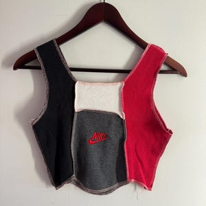 May include: A patchwork crop top in black, white, gray, and red. The top features a square neckline and is stitched together with visible seams. The Nike logo is embroidered in red on the gray panel. The top is hanging on a wooden hanger.