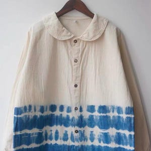 May include: Cream-coloured button-down shirt with a rounded collar and long sleeves. The bottom section of the shirt has a horizontal tie-dye pattern in blue hues. The shirt is hanging on a wooden hanger.