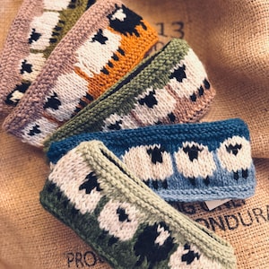 May include: A collection of four knitted headbands featuring a repeating pattern of white and black sheep. The headbands are displayed in various colorways including olive green, mustard yellow, light brown, and blue, each with a textured knit finish.