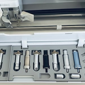 Könnte beinhalten: Eine weiße Schneidemaschine mit einer Auswahl an silbernen und schwarzen Schneidklingen und Werkzeugen. Die Werkzeuge sind in einer grauen Ablage angeordnet, mit einer Vielzahl von Formen und Größen. Der obere Teil der Maschine ist sichtbar.