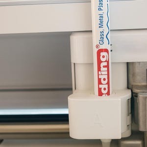 Könnte beinhalten: Ein weißer Edding-Marker mit roter Schrift und dem Text "Glass, Metal, Plastic" ist in eine weiße Maschinenkomponente eingesetzt. Die Komponente hat den Buchstaben "A" aufgedruckt. Der Hintergrund ist weiß.