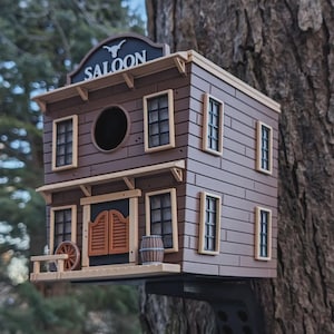 Könnte beinhalten: Ein braunes und beigefarbenes Vogelhaus aus Holz in Form eines Saloons, an einem Baum befestigt. Das Vogelhaus hat ein Schild mit der Aufschrift "SALOON" und dekorative Elemente wie ein Fass und ein Wagenrad.