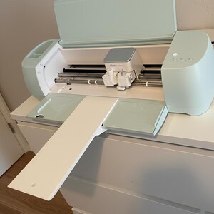 Könnte beinhalten: Eine mintgrüne und weiße Cricut-Schneidemaschine mit offenem Deckel und ausgezogenem Fach. Die Maschine hat einen silbernen Deckel und weiße Schneidköpfe. Sie steht auf einer weißen Kommode.