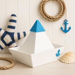 Könnte beinhalten: Ein weißer, bootförmiger Behälter mit einem blauen, dreieckigen Deckel. Das Boot ist an der Seite mit einem kleinen blauen Ankeremblem versehen. Die Szene umfasst einen gestrickten blau-weißen Seestern, ein aufgerolltes Seil, einen hängenden blauen Anker und eine Muschel.