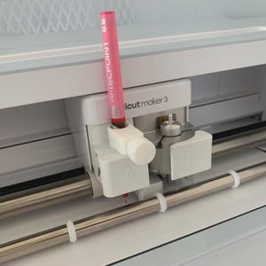 Könnte beinhalten: Eine weiße Cricut Maker 3 Schneidemaschine mit einem Doppelschlittensystem und den mit A und B gekennzeichneten Steckplätzen. Ein rosa Hi-Tecpoint 0.5 Stift steckt in der weißen Halterung an Steckplatz A über einer horizontalen Metallschiene.