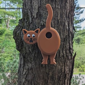 Könnte beinhalten: Braune Katzen-Dekoration mit Hintern und Kopf, an einem Baum befestigt. Das Hinterteil der Katze hat eine kreisförmige Öffnung, und der Kopf hat ein niedliches Gesicht mit Schnurrhaaren. Die Dekoration besteht aus einem festen Material.