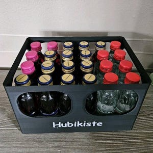 Könnte beinhalten: Eine schwarze Holzkiste gefüllt mit kleinen Glasflaschen mit bunten Verschlüssen. Auf der Kiste steht in weißer Schrift das Wort "Hubikiste". Die Flaschen haben rote, rosa und blaue Verschlüsse.
