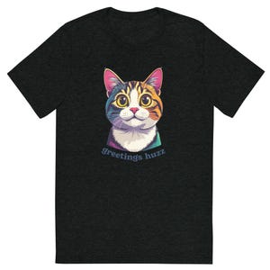 May include: Dark gray t-shirt featuring a colorful cartoon cat face with the text "Greetings huzz". The cat has a white muzzle, orange and brown fur, and pink ears. The shirt is made of a soft material.