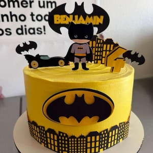 以下が含まれることがあります： バットマンをテーマにした黄色いケーキ。ケーキには黒いバットマンのロゴ、街並みのボーダー、バットマンのフィギュアが描かれています。ケーキの上にはバットマンのロゴと「Benjamin」という名前が飾られています。