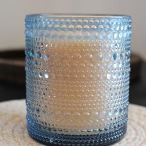 Peut inclure: Un bougeoir en verre bleu clair avec un motif texturé en forme de bulles. La bougie à l'intérieur est de couleur beige clair. Le bougeoir repose sur un dessous de verre blanc tissé.