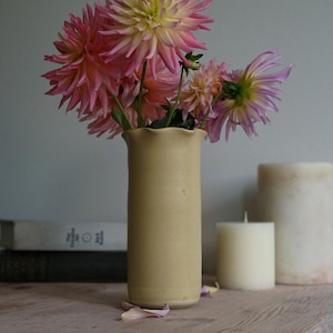 May include: A tall, cylindrical, matte beige ceramic vase with a gently scalloped rim. The vase holds a bouquet of vibrant pink and yellow dahlia flowers with long, slender petals. The vase has a smooth, uniform texture and a simple, clean design.
