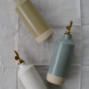 Op de afbeelding: Drie keramische olieflessen in olijfgroen, zeegroen en gespikkeld wit, elk met een goudkleurige tuit. De flessen hebben een lichtbeige basis en zijn gerangschikt op een getextureerd, lichtgrijs oppervlak. Ideaal voor in de keuken.