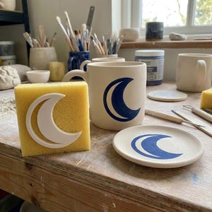 May include: A ceramic mug and a small round ceramic plate, both featuring a dark blue crescent moon design. A yellow square sponge with a white crescent moon cutout sits in the foreground, alongside various pottery tools and supplies.