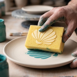Può includere: Una spugna gialla con un disegno a conchiglia viene utilizzata per applicare vernice turchese a un piatto di ceramica bianco. Il piatto ha un disegno a conchiglia abbinato. L'immagine mostra strumenti e forniture per la ceramica.