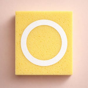 May include: A square, yellow sponge with a white circular design in the centre. The sponge has a textured surface and is set against a light pink background. The white circle is a contrasting element.