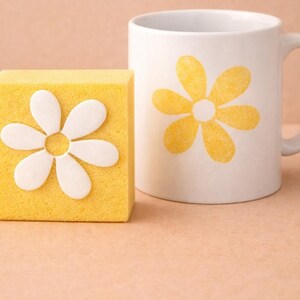 Puede incluir: Un sello de esponja amarilla con un diseño de margarita blanca junto a una taza blanca con una impresión de margarita amarilla. El fondo es de un tono suave y neutro. El texto "HmcStamps" es visible.