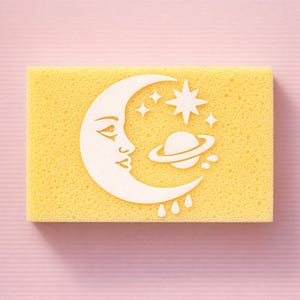 May include: A yellow sponge with a white celestial design featuring a crescent moon, stars, and a planet with rings. The design is centered on the rectangular sponge, creating a whimsical and decorative element.