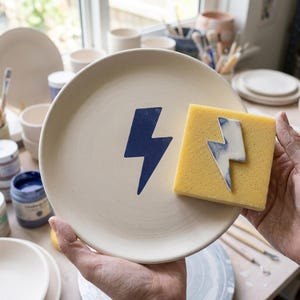 Op de afbeelding: Een rond, gebroken wit keramieken bord met een donkerblauw bliksemschichtontwerp in het midden. Een vierkante gele spons met een bijpassende bliksemschicht-uitsparing wordt tegen het bord gehouden, wat het sjabloon voor het ontwerp toont.