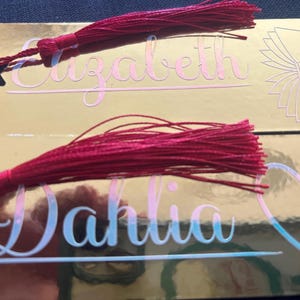 Könnte beinhalten: Zwei rechteckige, goldfarbene Lesezeichen mit reflektierender Oberfläche. Das obere Lesezeichen zeigt den Namen Elizabeth in rosa Schreibschrift und ein Buch-Symbol. Das untere zeigt den Namen Dahlia in blauer Schreibschrift und ein Herz. Beide haben rote Quasten.