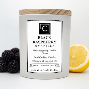 May include: A white candle with a wooden lid, labeled "Black Raspberry & Vanilla." The label includes the scent description, "Black Raspberry, Vanilla, Citrus," and states it is hand-crafted with essential oils. A sliced lemon and blackberries are in the background.