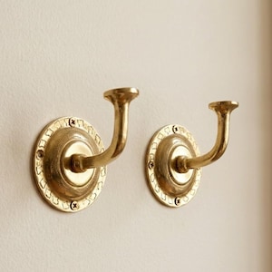 May include: Two brass wall hooks. Each hook has a circular base with decorative detailing and a curved arm ending in a rounded top. The hooks are mounted on a light-colored wall, providing a classic and elegant storage solution.