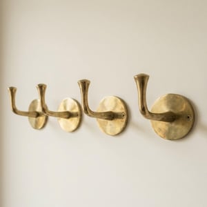 May include: Four brass wall hooks with a curved design are mounted on a white wall. Each hook has a circular base and a curved arm, perfect for hanging coats, hats, or other items. The hooks are made of a warm, golden-toned metal.