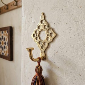 May include: A gold-toned brass wall hook featuring an intricate diamond-shaped backplate with cut-out circular patterns and engraved detailing. A curved hook extends from the base, holding a multi-colored yarn tassel with orange, blue, and brown threads.