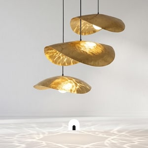 May include: Three modern pendant lights with a unique, organic design. Each light features a golden, leaf-like shade with a textured surface, suspended from a black cord. The lights are illuminated, casting a warm glow and intricate patterns on the surrounding white walls and floor.