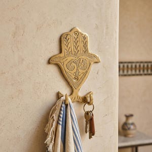 May include: A gold-toned brass wall hook shaped like a Hamsa hand with intricate engraved patterns. The hook features two prongs at the bottom, one holding a folded blue and white striped fabric and the other holding a metal keyring with keys.